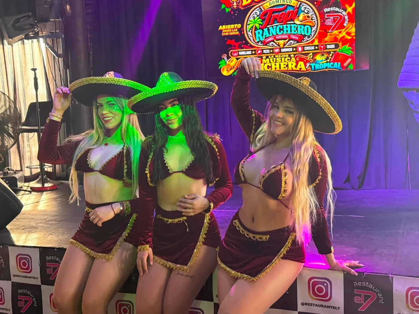 Las tres integrantes de Reinas Latinas con sombreros de charra y trajes granate frente a un escenario con luz verde y violeta