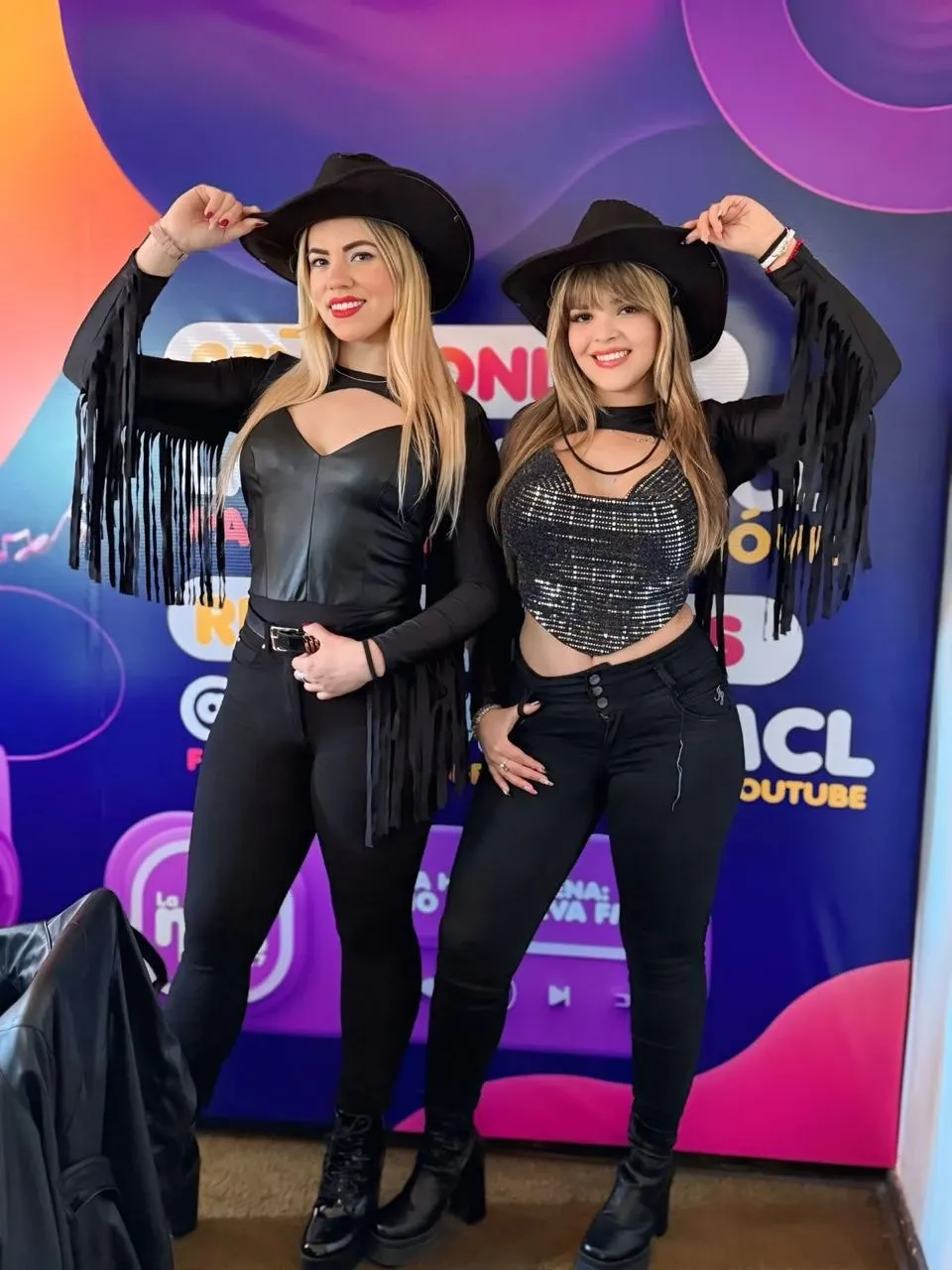 Dos integrantes de Reinas Latinas con sombreros negros de fleco frente al telón de Radio La Nueva FM