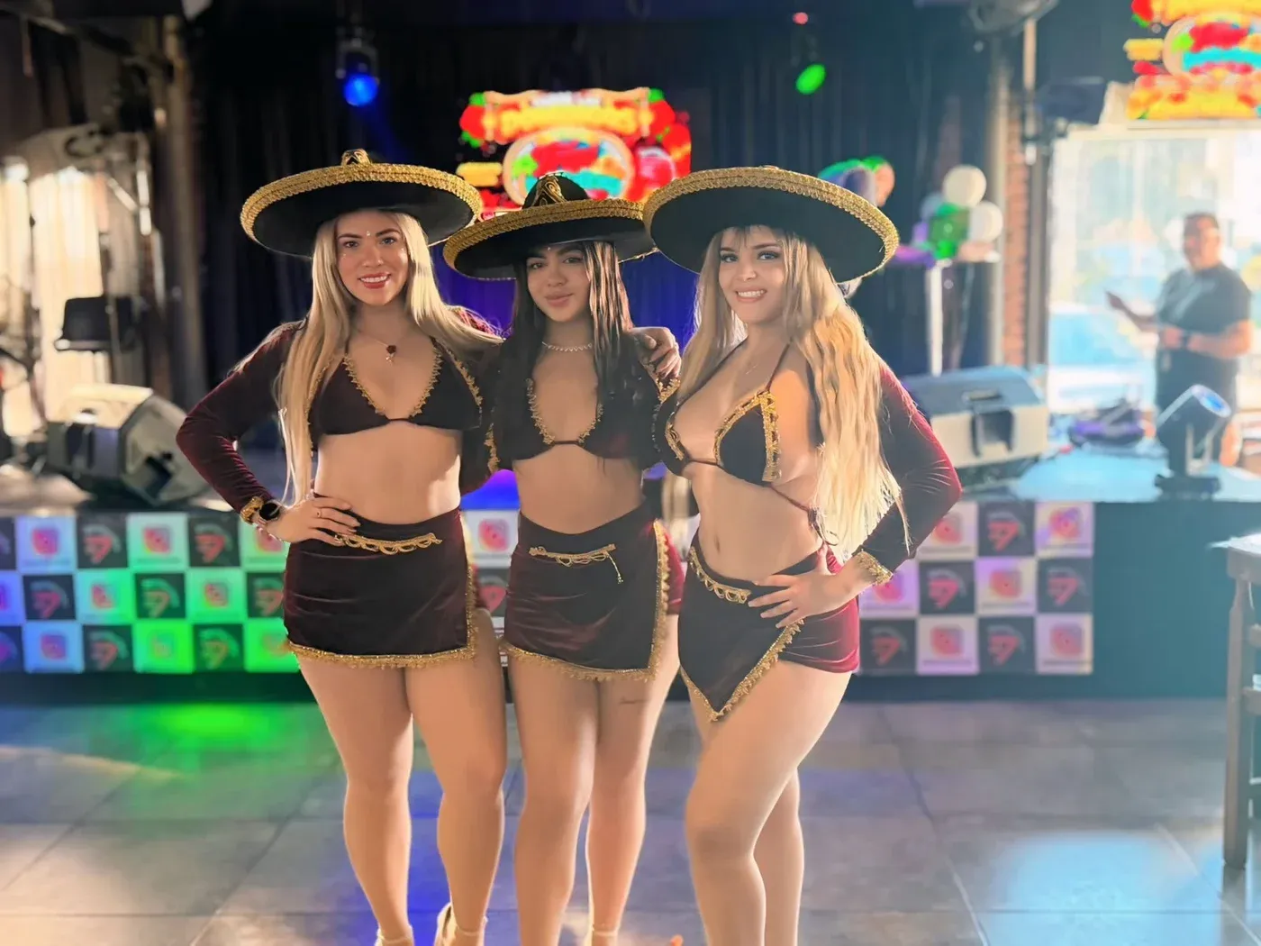 Las tres integrantes de Reinas Latinas con sombreros de charra y trajes granate frente a un escenario iluminado