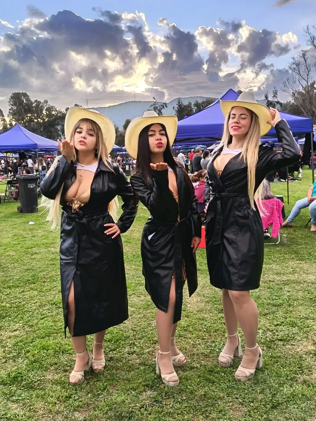 Las tres integrantes de Reinas Latinas con sombreros claros y abrigos negros lanzan besos a la cámara en una feria al aire libre con toldos azules