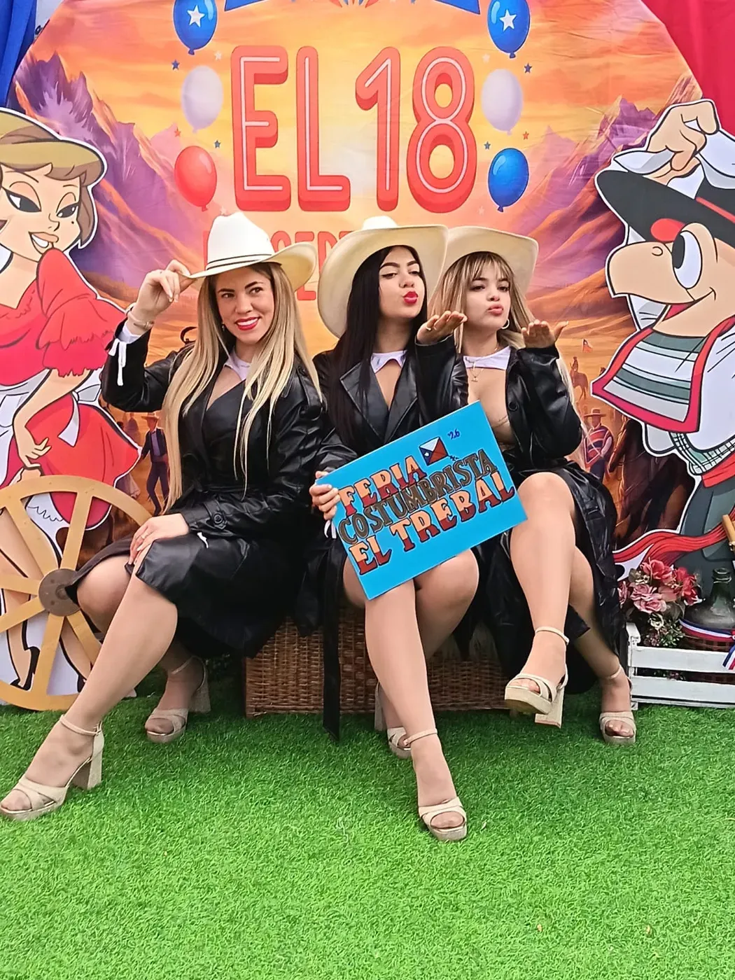 Las tres integrantes de Reinas Latinas con sombreros claros y abrigos negros, sentadas en un photocall del 18 con un cartel de la Feria Costumbrista El Trebal