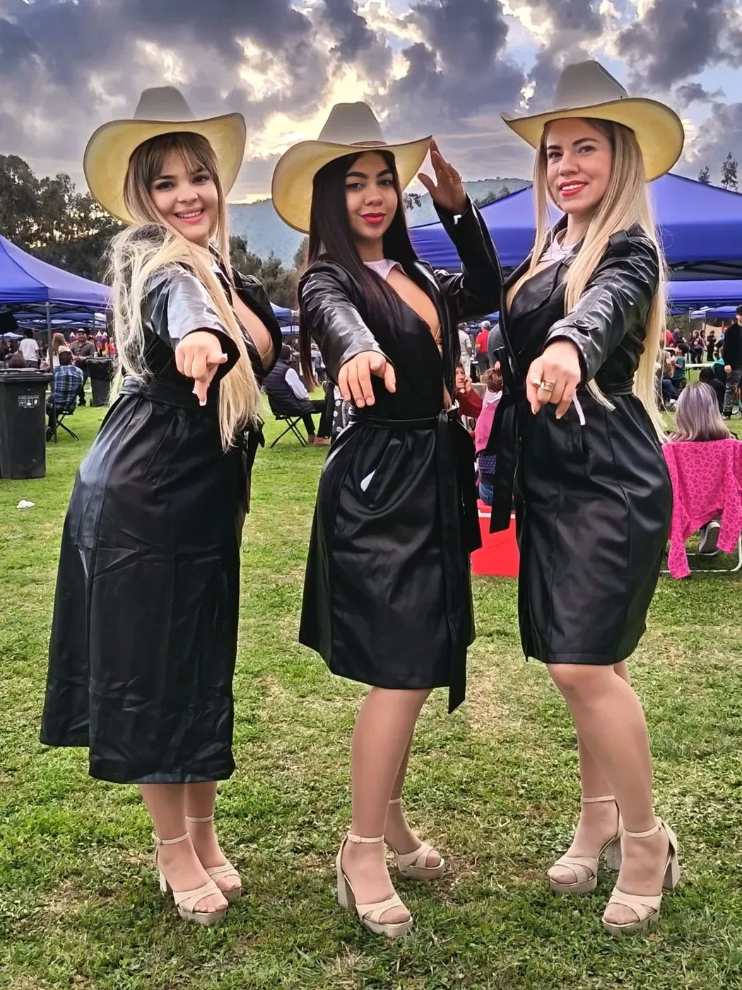 Las tres integrantes de Reinas Latinas con sombreros claros y abrigos negros apuntan a la cámara sobre el pasto de una feria al aire libre